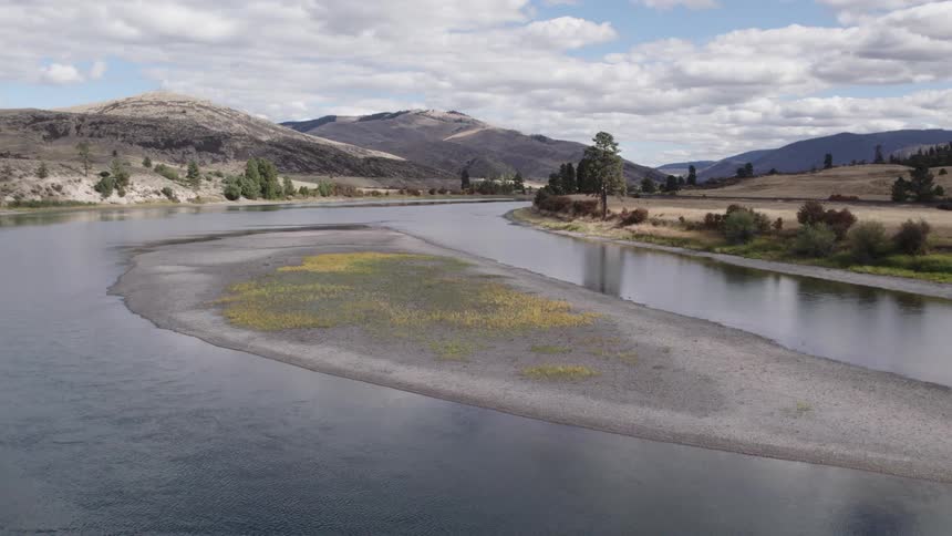 A crescentshaped islet with sparse grass on a slowly flowing river sits under a cloudy sky near sparse vegetation and gray mountains