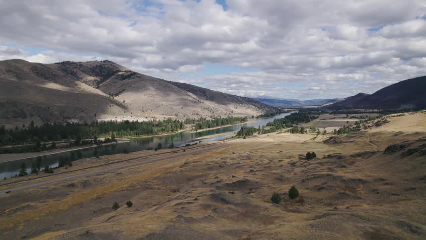 Conifer trees line river passing through arid scrubby terrain and mountains