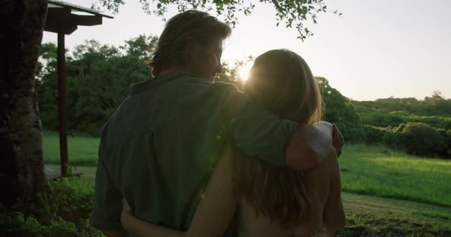 Man with shirt holds and kisses woman with blonde wavy hair next to country house at sunset in the countryside