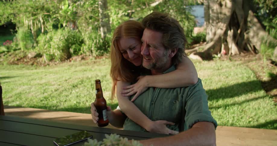 Woman hands man sitting at picnic table a beer then back hugs him