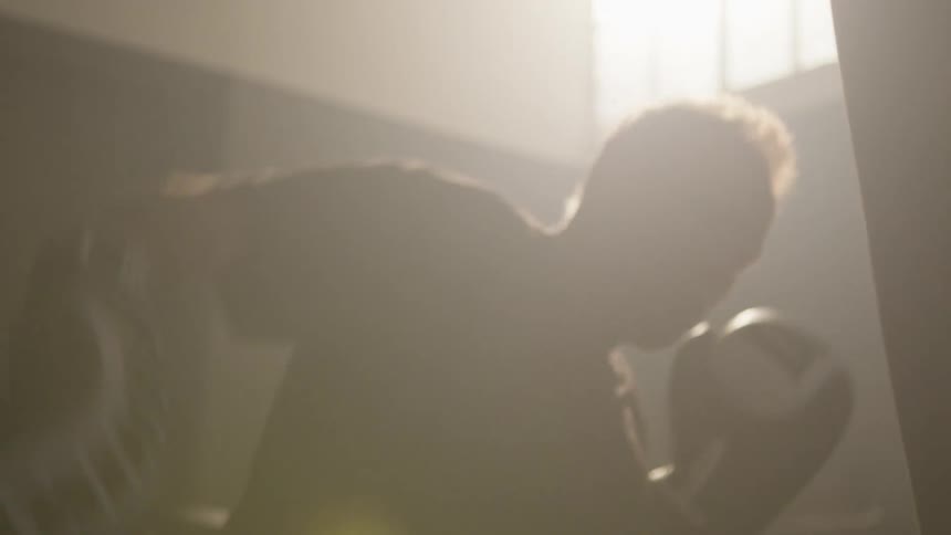Sunlight filters through a window over a man boxing with a punching bag on a dimly lit gym at morning