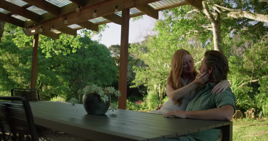 A woman caresses her husbands face while she sits on his lap at their patio table