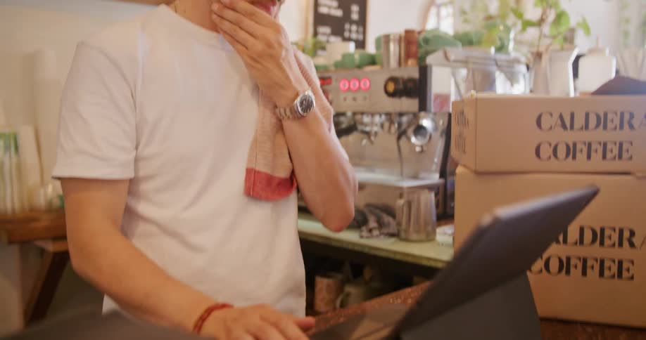 Man with moustache and white t shirt types on tablet keyboard behind the cafe counter at cafe