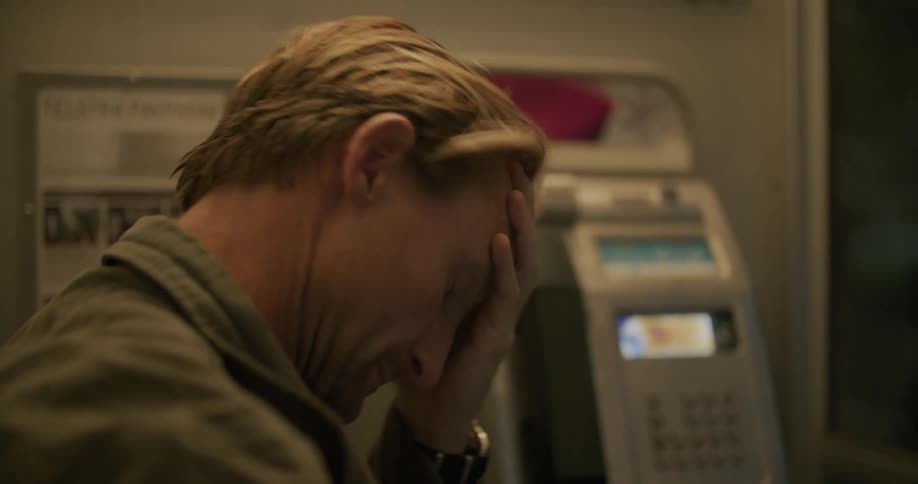 A man rubs his head after using a public phone and puts on his hat before leaving