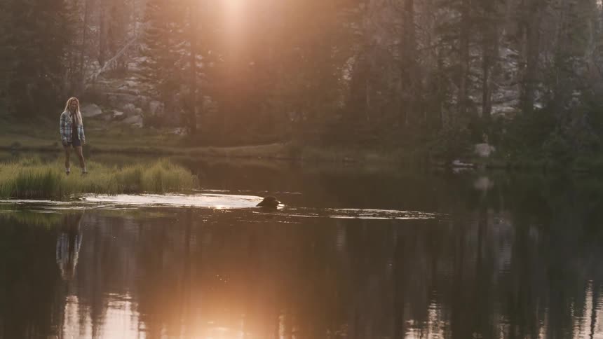 A man sits outside a tent while a woman throws a ball in to a river and a dog jumps in and swims to retrieve it