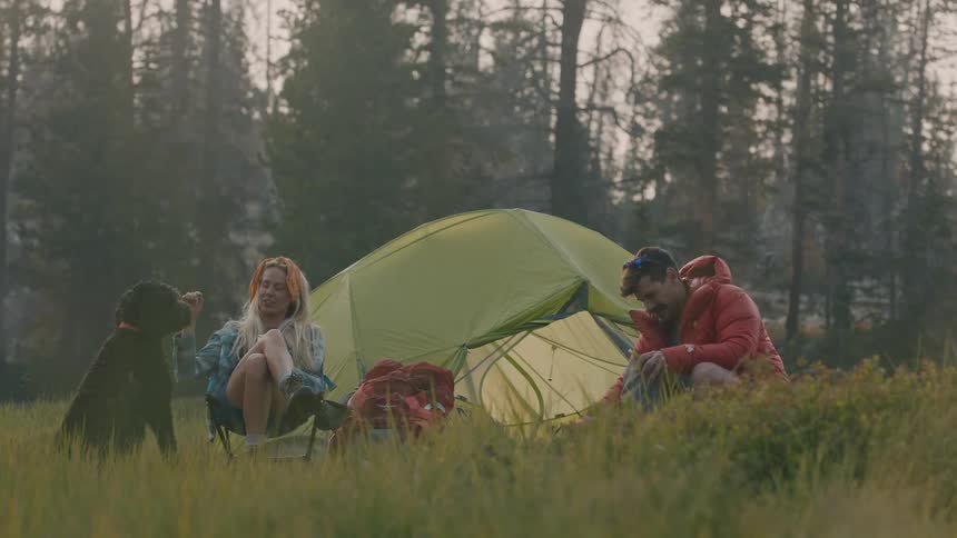 A man standing in front of a tent in a forest area bends down and takes a tripod out of a bag as a woman sits next to a dog and starts petting it