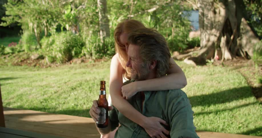 Woman hands man a beer bottle and then back hugs him as he sits at picnic table near trees allowing glimpse of water
