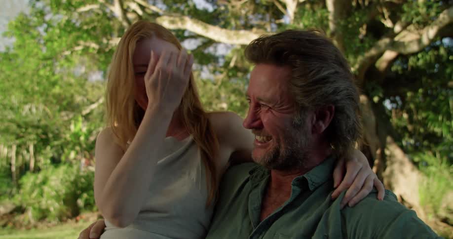 Man with beard and blue shirt holds woman with red hair and grey vest sitting in garden laughing and lovingly looking into each others eyes