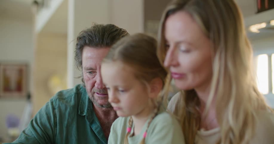 Father with beard blue shirt daughter with braids and blonde mother wearing brown dress use and look at tablet in kitchen at home