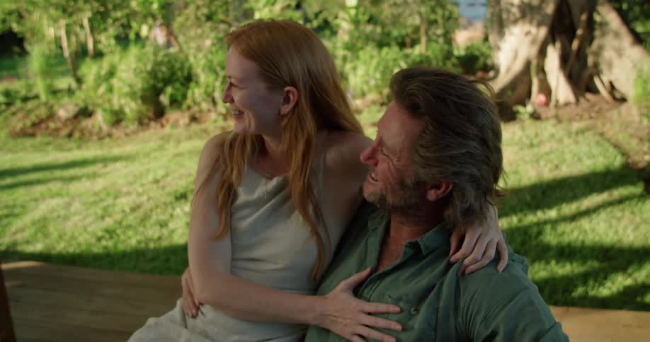 Man with beard and blue shirt holds woman with red hair and grey vest sitting in garden lovingly looking into each others eyes and kissing