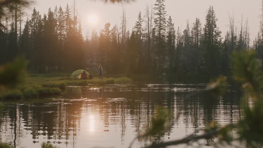 A man and woman in front of a tent pitched on the shore look at the water