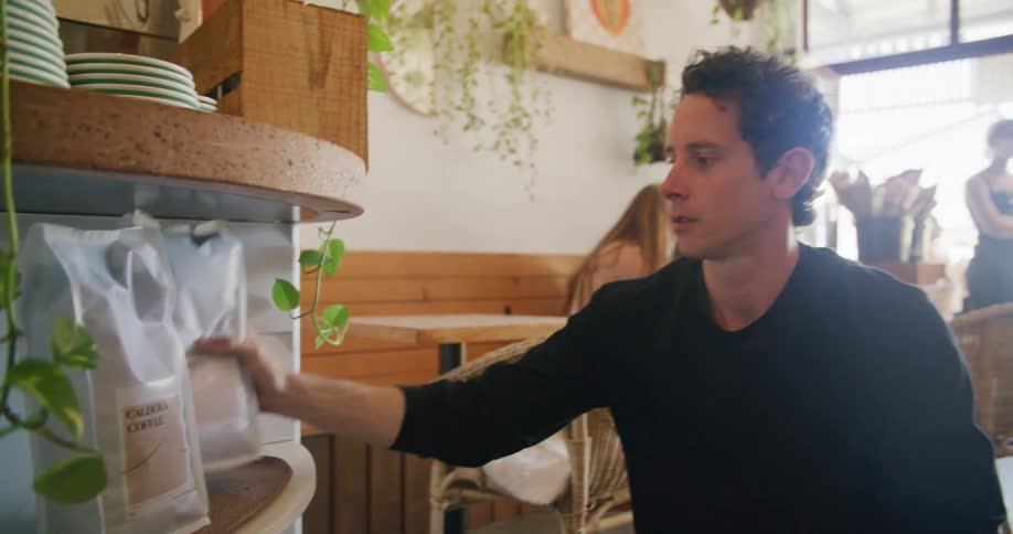 A man unpacks bags of coffee from a box and places them on a shelf in a cafe
