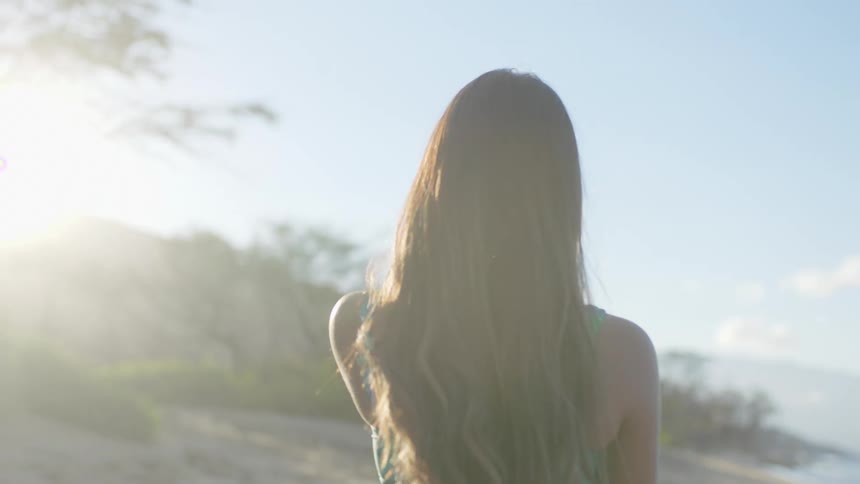 Woman with brown wavy hair and turquoise swimming costume walks on the beach next to sea