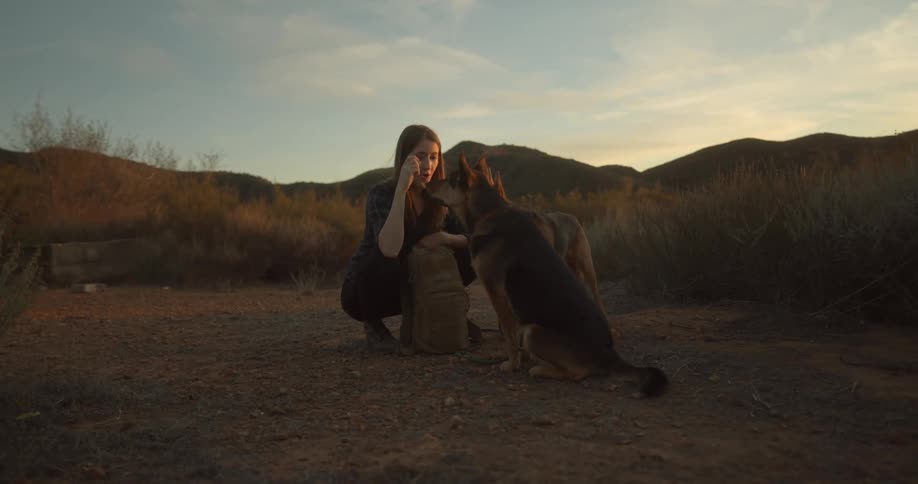 A woman feeds and pets a pair of dogs while kneeling in the countryside during the day