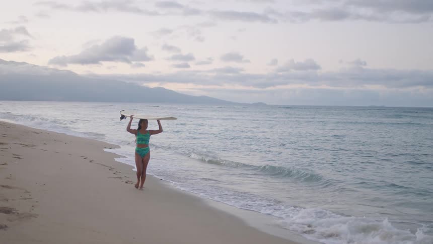 Young woman wearing a swimsuit walks on the beach with a surfboard over her head at sunset