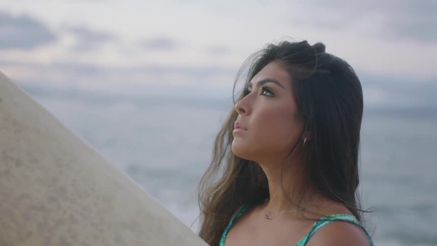 A woman standing on the shore runs her fingers through her hair and turns her head and smiles