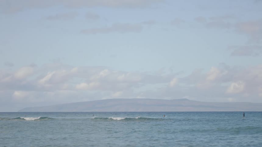 Waves are rolling in as a couple of people are in the water and a mountain is in the distance