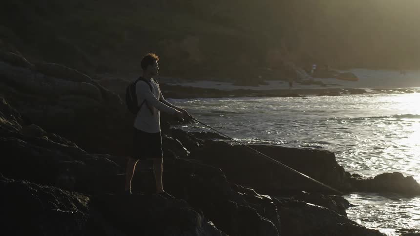 A man stands on a rocky coastline and fishes