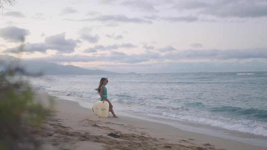 A young woman sits on her surfboard on the beach and watches the waves roll in