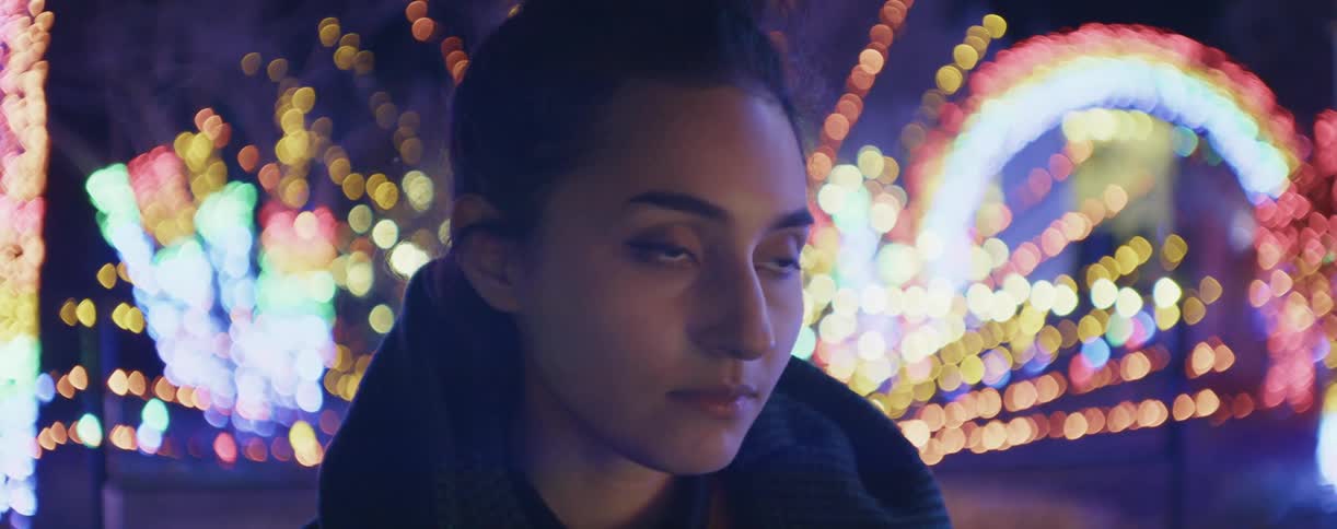 A teenager is standing outside looking around with bright flashing colored lights in the distance