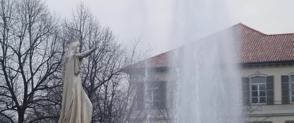 Fountain decorated with statue standing near red roofed house and leafless trees shoots water into the air
