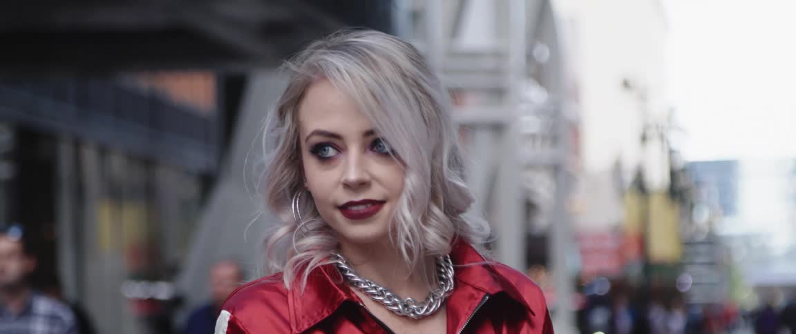 Woman with makeup gray hair hoop earrings and red jacket looks around and raises her hand smiling in the city street