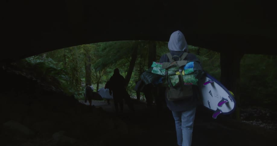 Group of surfers hiking on the forest walk under a bridge carrying surfboards