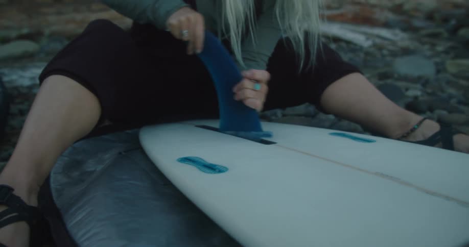 Woman sitting on the ground of a forest gluing the fin on the bottom of a surfboard at morning