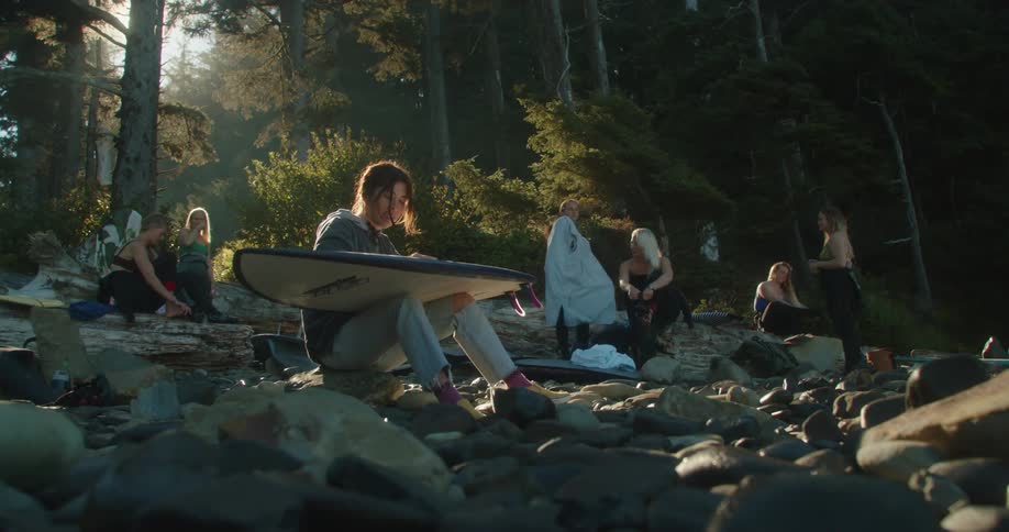 Group of female friends with surfboards sit around stone strewn area prepping