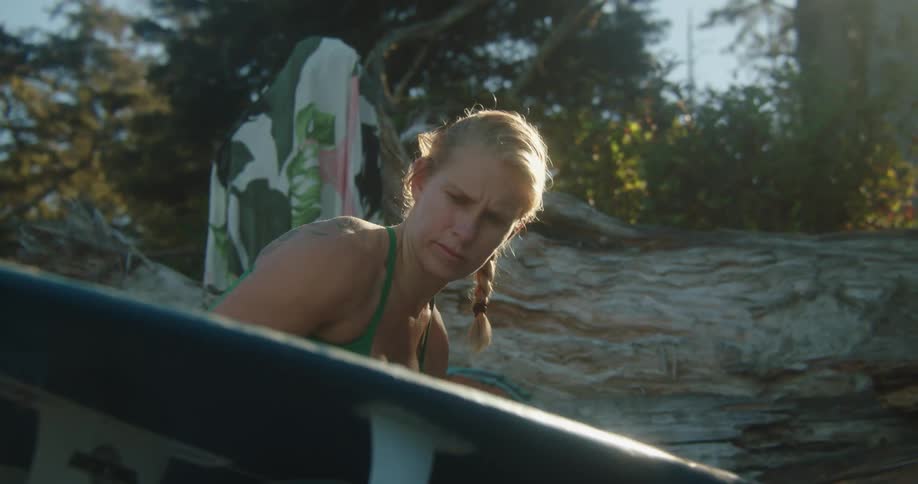 Blonde woman waxes board sitting on ground with trees in background