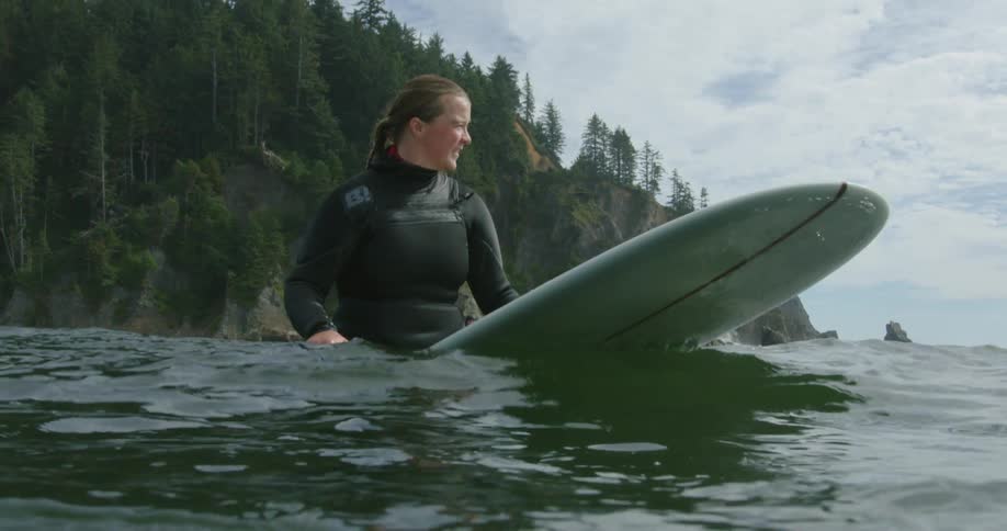 A woman sitting on a surfboard bobbing up and down with the gentle waves near a tree covered coast