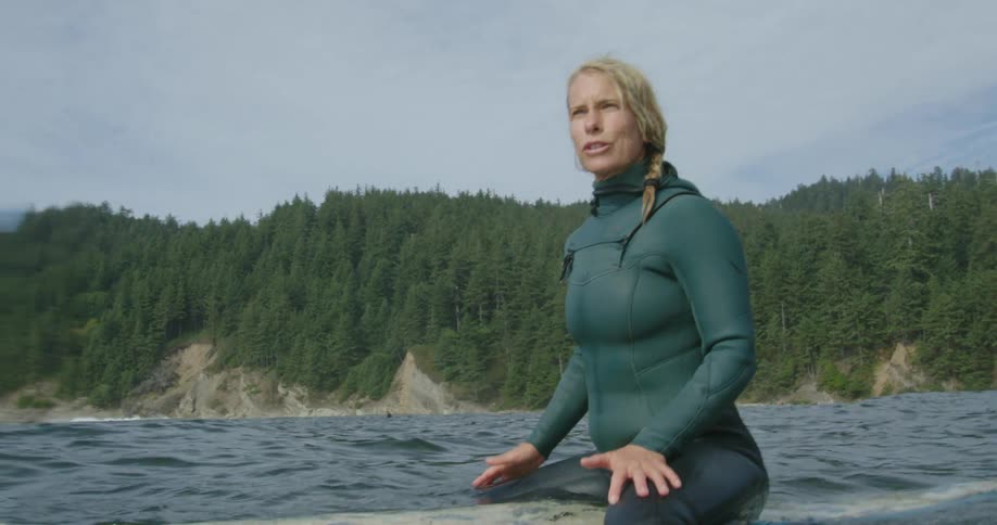 Blonde woman wearing a wet suit sitting on a surfboard floating on the ocean near the mountains
