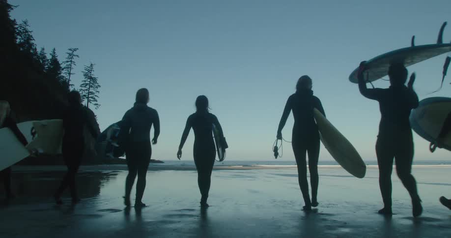 A group of people wearing wetsuits are carrying surfboards across a beach
