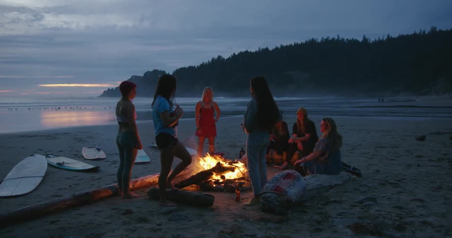 A group of women talk around a campfire on the beach at sunset while one woman plays the ukulele