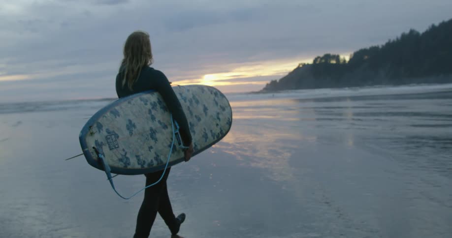 A surfer wearing a wetsuit carries their board toward the water at sunset
