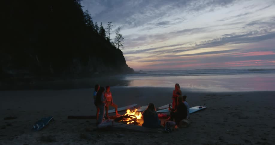 A group of female surfers stand are gathered around the campfire