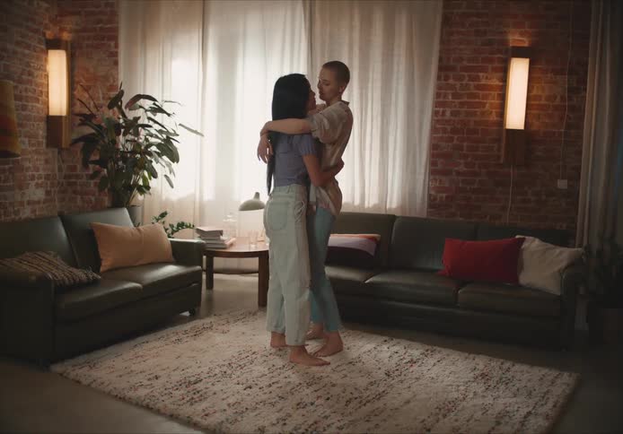 Short hair woman with shirt and jeans holds and dances with woman with long straight hair purple shirt and jeans in living room at home