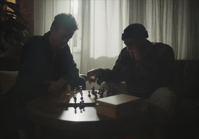 A pair of men laugh while playing chess together in a dimly lit room during the day