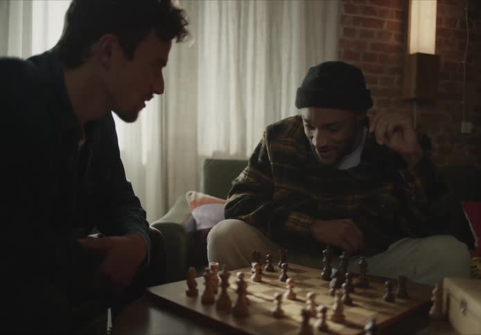 Two friends laugh and talk as they play chess at home