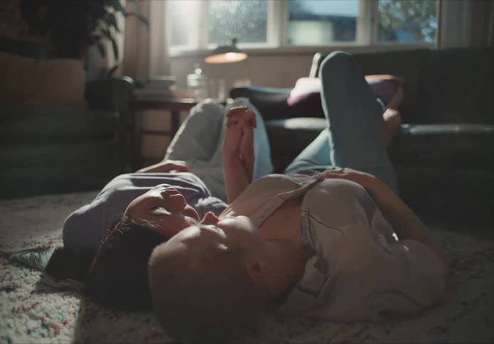 Two women lie on a living room floor holding hands