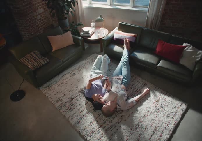 Two women lie on their backs on living room rug talking about contents of mobile phone