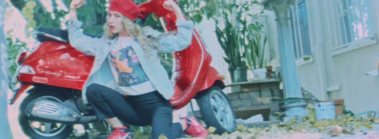 Woman crouches and poses in front of red scooter near house