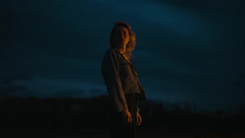 A woman is standing outside alone at night as her blonde hair moves in the breeze