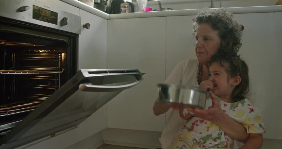 A little girl sits in her grandmothers knee on the kitchen floor and the grandmother opens the oven and puts a pan in
