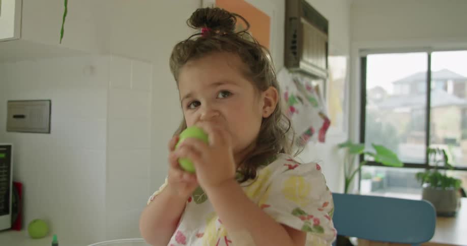 A little girl sitting on the kitchen counter takes a bite of a green apple