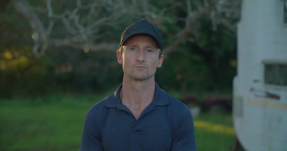 A man wearing a dark colored cap is standing outside with trees behind him