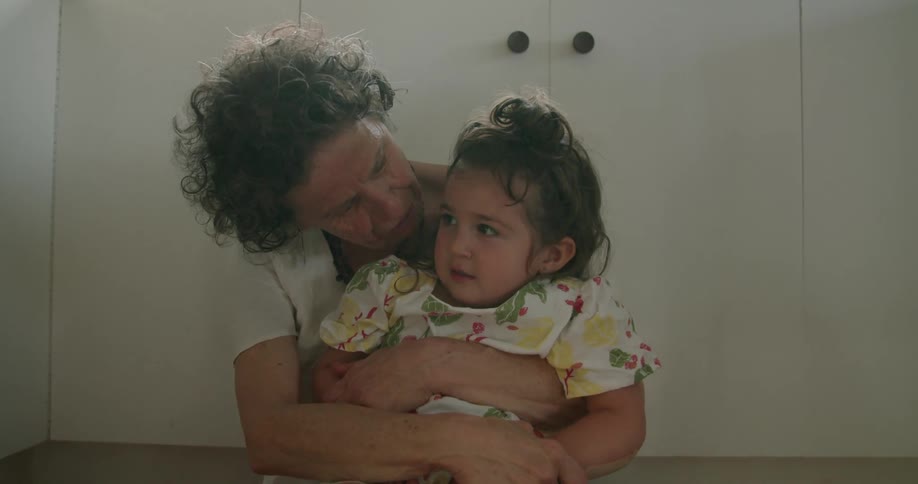 A grandmother is sitting on the kitchen floor with her arms around her granddaughter as they talk