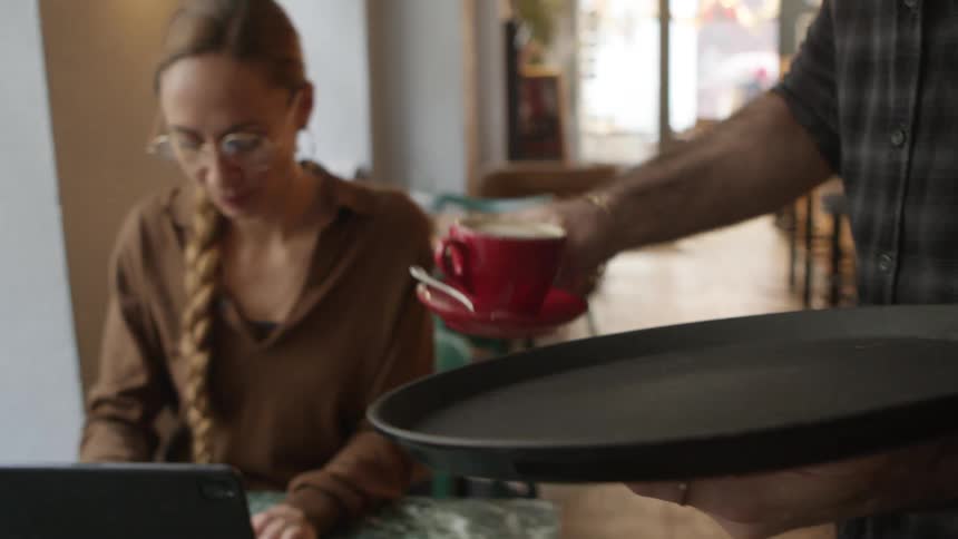 A waiter brings a mug of coffee to a woman working on her laptop in a cafe