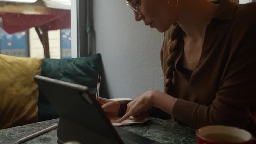 A woman sits at a desk in an office and writes in a notebook in front of a tablet