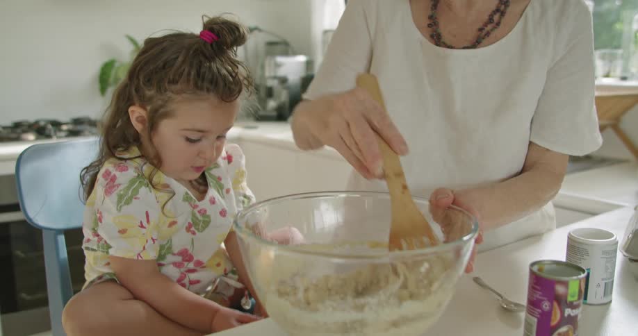 A young girl is in the kitchen with her grandmother watching her stir a mixture in a bowl with a wooden spoon